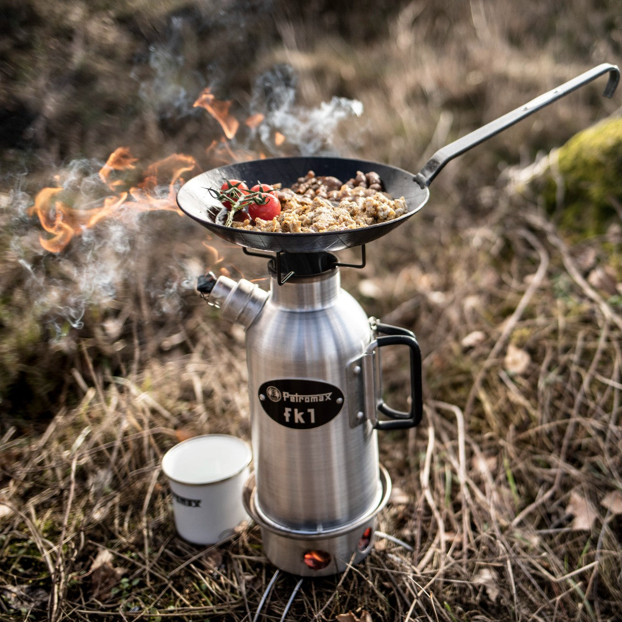Outdoor camping stove setup with a compact stainless steel kettle and black frying pan cooking food with tomatoes over an open flame, featured in Kitchen & Food, Cookware, Cast Iron, and Best Sellers collections.
