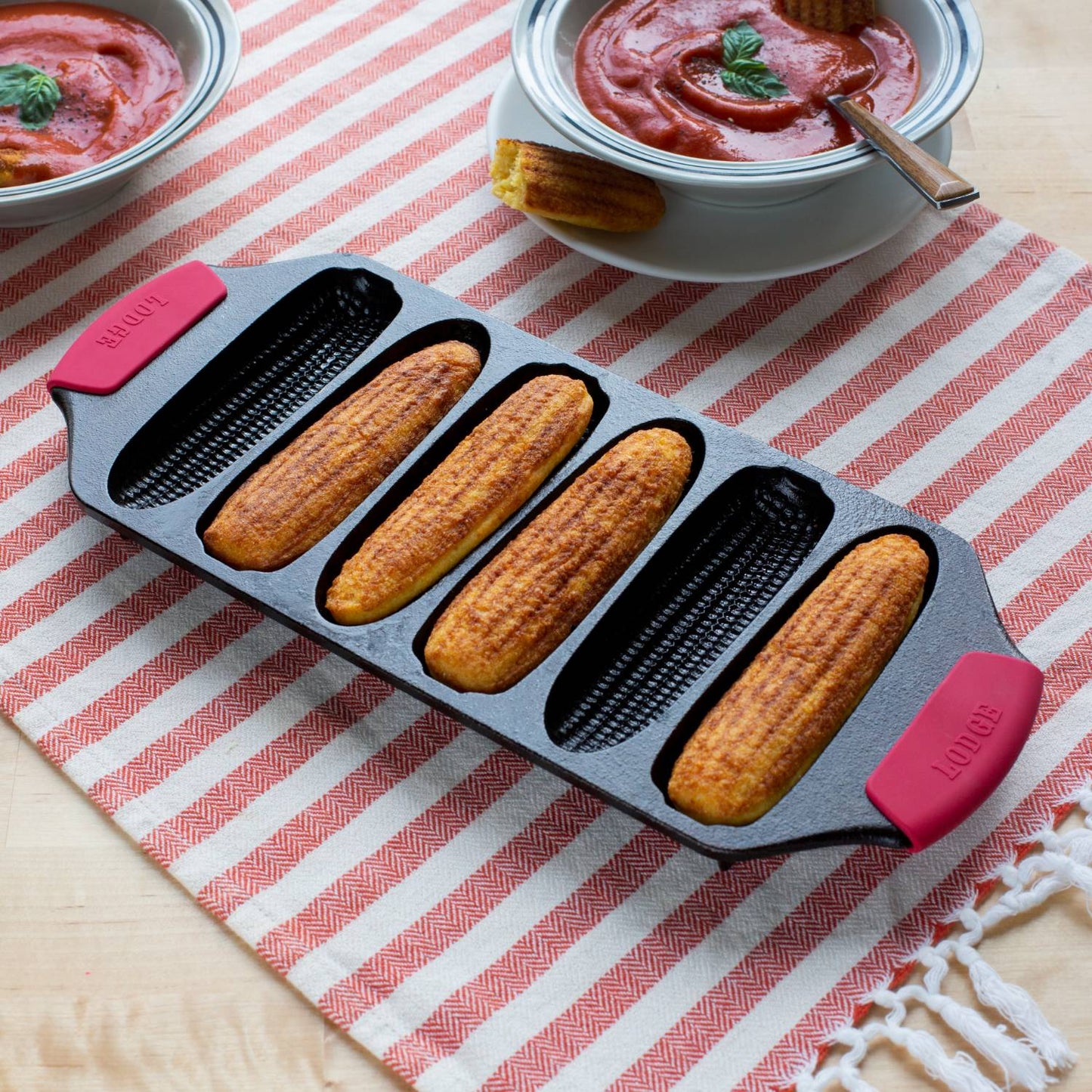 Seasoned Lodge cast iron cornstick pan with six golden cornbread sticks, featuring dual red silicone handles and angled feet, displayed on a striped cloth with bowls of tomato soup, ideal for Kitchen & Food, Cookware, Cast Iron, USA Made, Best Sellers, and Promo Eligible collections.