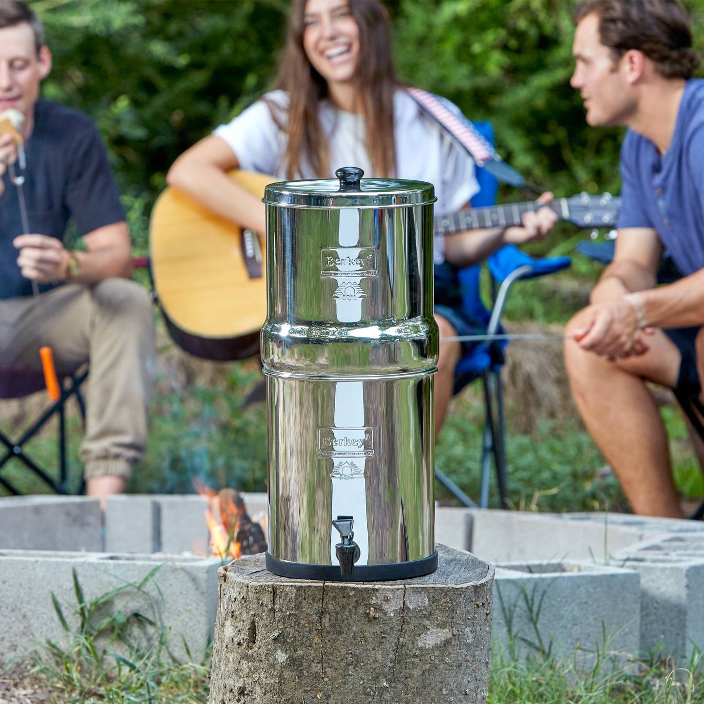 Highly polished stainless steel water filtration system with black spigot and stacked chambers, positioned outdoors on a tree stump near a campfire, surrounded by people enjoying music, suited for Farm, Garden & Tool, Appliances, and Berkey Water Filters collections.