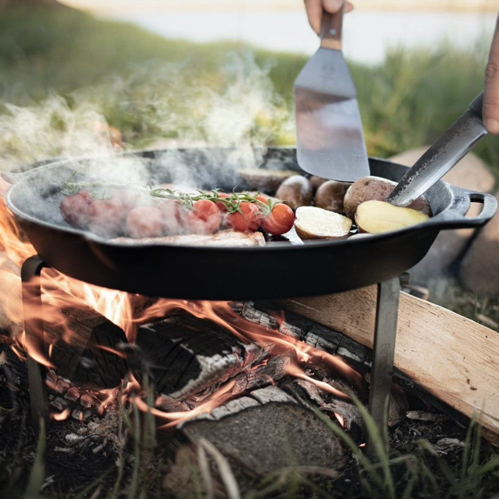 Outdoor cooking over an open wood fire with a cast iron pan filled with vegetables and meat being turned using a flexible spatula with a wooden handle, ideal for grilling and kitchen gadgets collections.