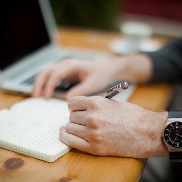 Close-up of a person writing notes with a silver pen in a notebook on a wooden table, with a blurred laptop in the background, evoking focused productivity for Best Sellers and P3 Testing collections.