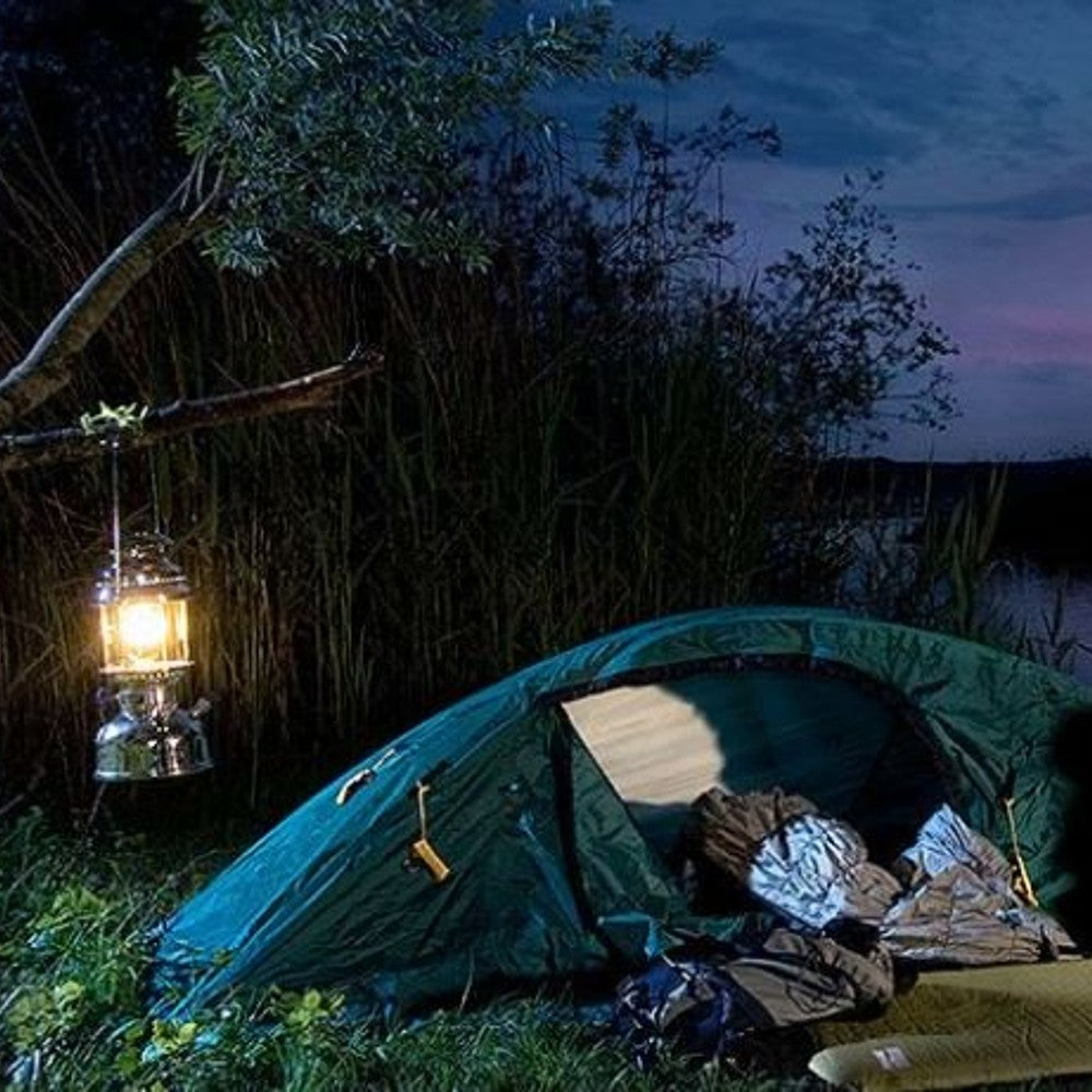 Glowing lantern hanging from tree branch beside a green camping tent pitched near dense reeds at dusk, depicting an outdoor scene fitting Lamps & Lighting, Parts and Accessories, Other Parts, Best Sellers, Promo Eligible collections.