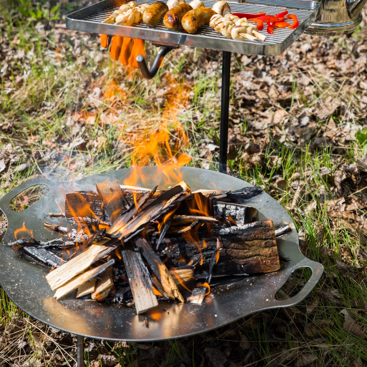 Outdoor fire bowl with a blazing wood fire beneath a raised grilling surface cooking sausages and skewers, perfect for camping, garden, and farm use in popular seasonal collections.