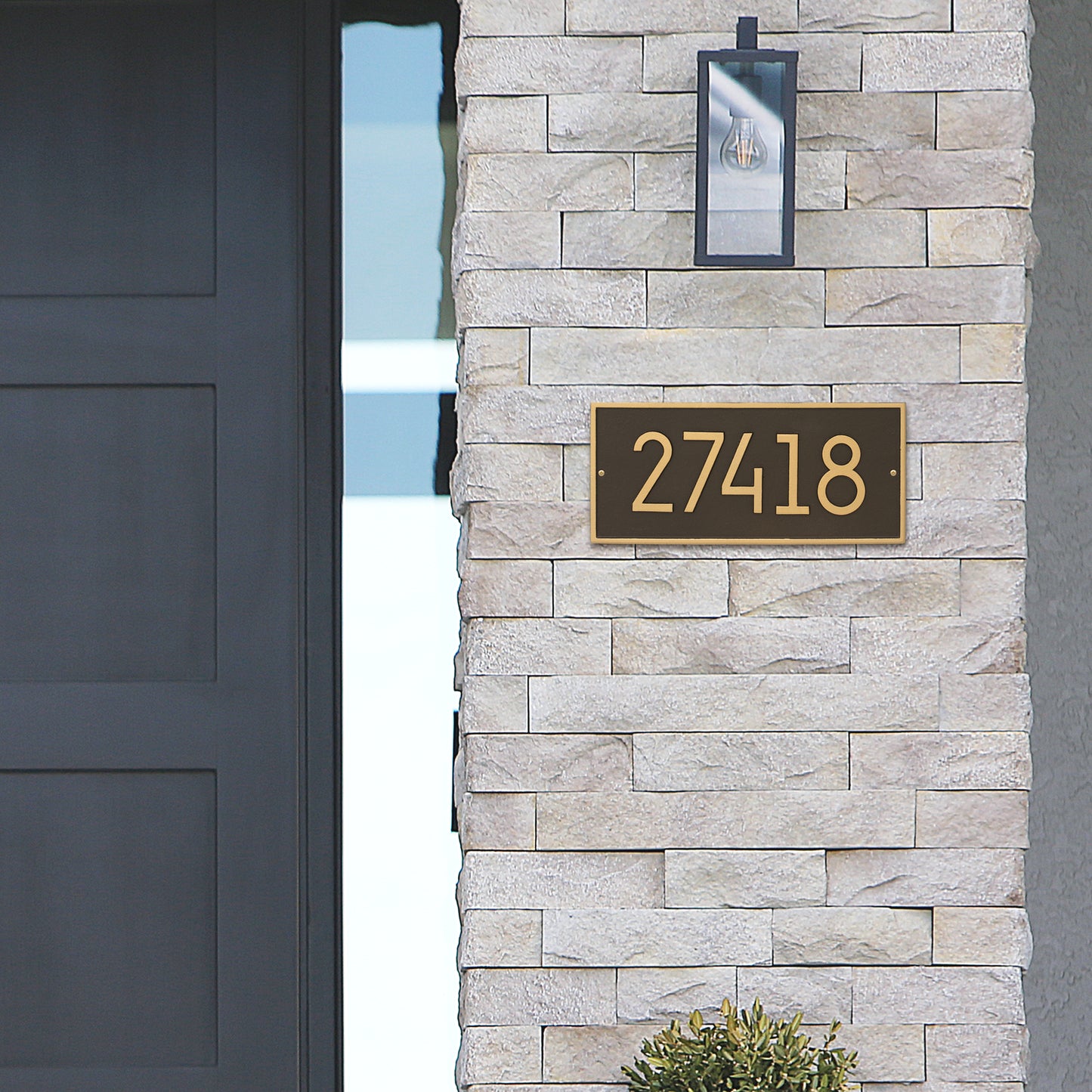 Modern custom address plaque in aged bronze with gold-tone numbers mounted on light-colored stone wall next to a door and outdoor lantern, handcrafted in USA, featured in Home & Decor, Mailboxes and Address Signs collections.