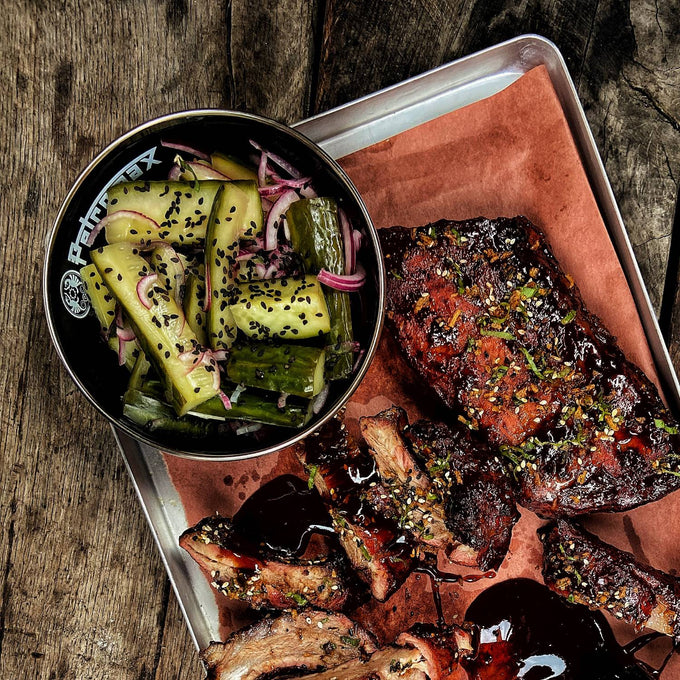 Black enamel bowl with white logo filled with seasoned pickles and onions, placed on a metal tray with grilled ribs, perfect for Farm, Garden & Tool, Kitchen & Food, Camping, Dinnerware, Best Sellers, and Promo Eligible collections.