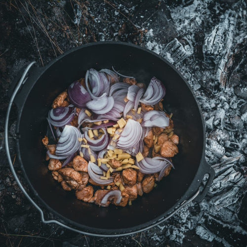 Petromax cast iron Dutch oven with flat base containing seasoned chicken, chopped onions, and garlic, cooking outdoors over glowing embers, featured in Kitchen & Food, Cookware, Cast Iron, Best Sellers, and Promo Eligible collections.