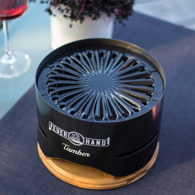 Sleek black tabletop charcoal grill with a distinctive radial top grate design, resting on a wooden base, set on an outdoor table, perfect for grilling in Farm, Garden & Tool and Camping collections.