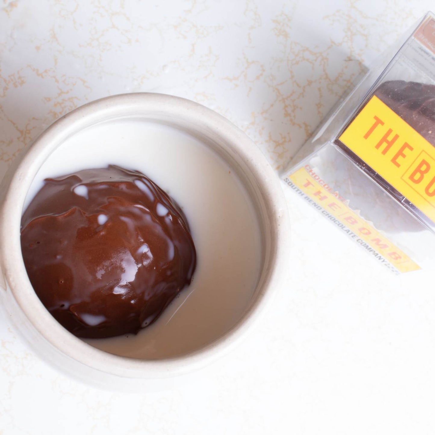 Handcrafted hot chocolate bomb with marshmallows melting in a white ceramic mug filled with warm milk, displayed beside clear packaging, ideal for Kitchen & Food, Snacks and Sweets, Beverages, USA Made, Best Sellers, and Promo Eligible collections.