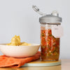 Clear wide-mouth jar with a silver fermentation lid and airlock filled with mixed vegetable ferment, labeled with a handwritten date tag, displayed next to a bowl of potato chips on an orange cloth, ideal for Kitchen & Food, Food Preservation, Fermenting and Pickling, USA Made, and Best Sellers collections.