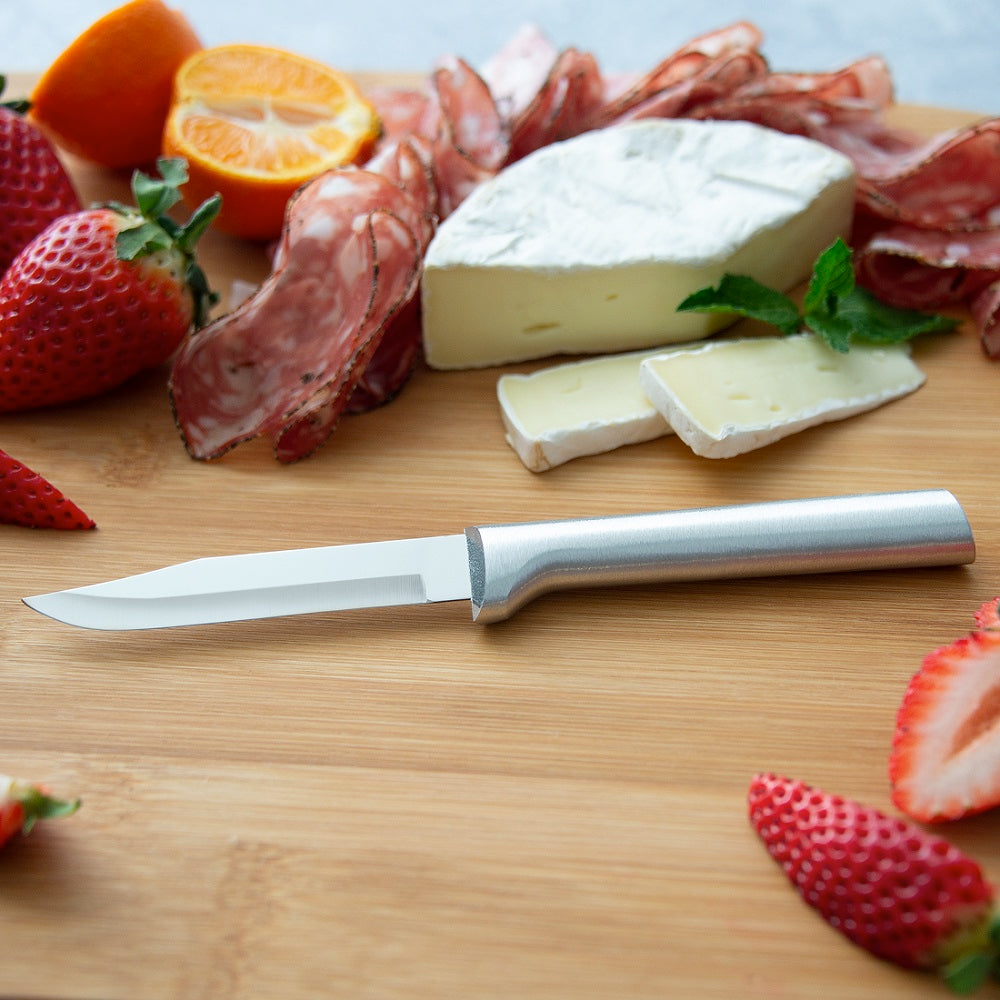 Sharp carbon stainless steel paring knife with a satin-finished cast aluminum handle, displayed on a wooden board with sliced strawberries, orange, cheese, and cured meats in a kitchen and food prep setting.