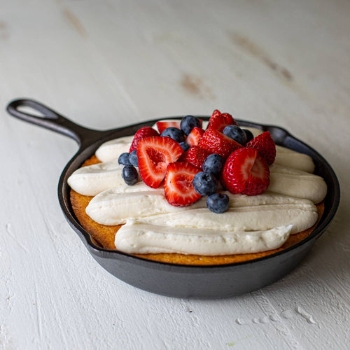 Pre-seasoned cast iron skillet holding a golden baked dessert topped with whipped cream and fresh strawberries and blueberries, featured in Kitchen & Food, Cookware, Cast Iron, Best Sellers, and Promo Eligible collections.