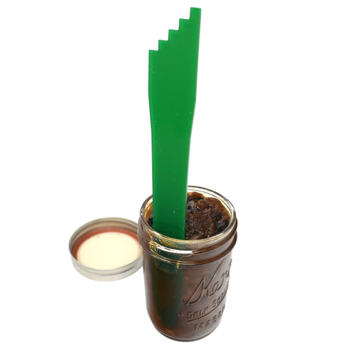 Green plastic canning bubble popper tool standing upright inside a glass jar filled with dark preserves, next to an open metal jar lid, for Kitchen & Food and Food Preservation collections.