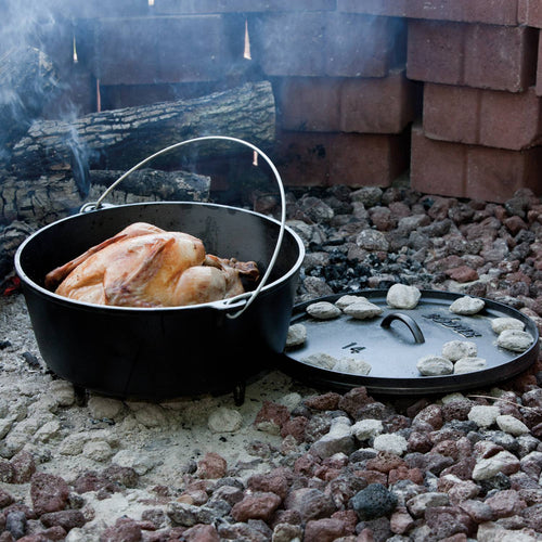 Handcrafted cast iron camp Dutch oven with built-in legs cooking a whole chicken outdoors over hot coals, designed for Farm, Garden & Tool, Camping, Cast Iron, USA Made, and Best Sellers collections.