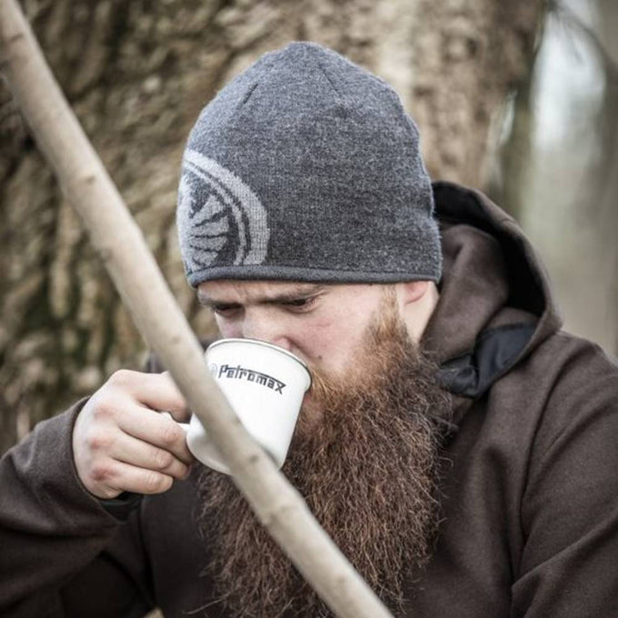 Man wearing a gray knit hat and brown hoodie sipping from a white enamel mug with black logo outdoors near a tree, suitable for Farm, Garden & Tool, Camping, and Brewing Coffee and Tea collections.