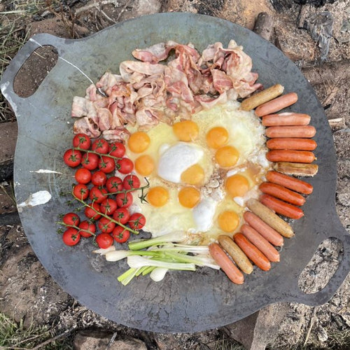Large steel cooking griddle with two handles sizzling eggs, bacon, sausage, cherry tomatoes, and green onions over an open fire, perfect for Farm, Garden & Tool, Camping, Best Sellers, and Promo Eligible collections.