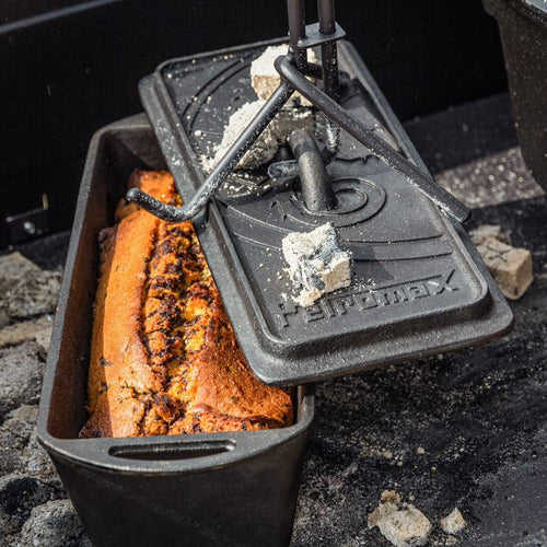 Cast iron loaf pan with freshly baked bread inside, next to a cast iron lid with ash and charcoal, demonstrated in outdoor cooking for camping and bread making supplies.
