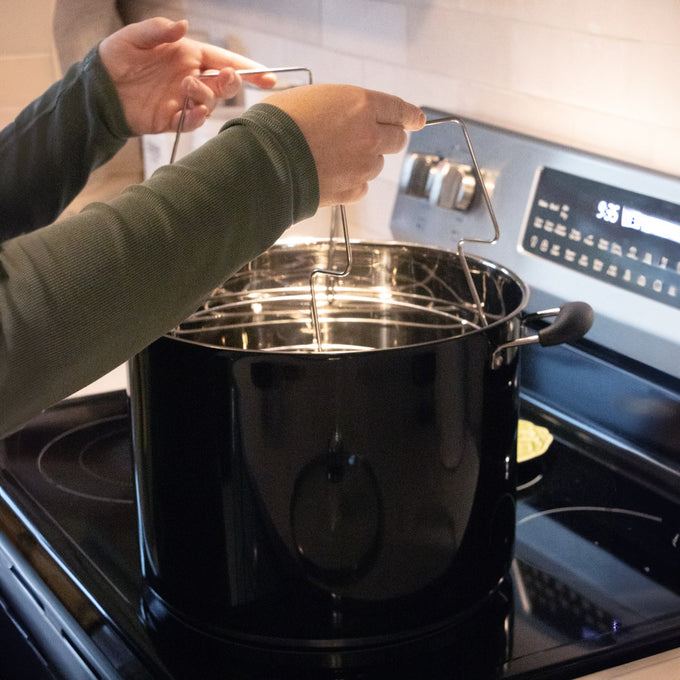 Person lifting a wire rack out of a glossy black 21.5-quart water bath canner on a stovetop, featured in Kitchen & Food, Canning, and Food Preservation collections.