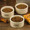 Three handcrafted natural stoneware pet bowls labeled "Kitty" and "DOG," filled with dry food, set on a rustic wood surface with pet treats nearby, highlighting USA made home and pet supplies.
