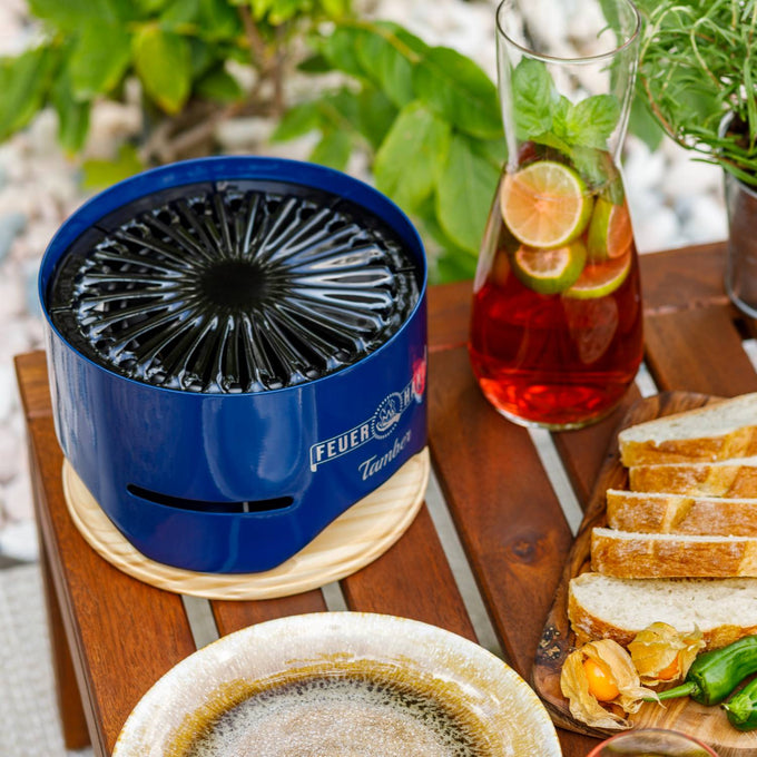 Compact blue tabletop charcoal grill with unique spiral top design resting on a wooden trivet, set on an outdoor wooden table with sliced bread and a glass pitcher of fruit-infused iced tea, featured in Farm, Garden & Tool and Grilling collections.