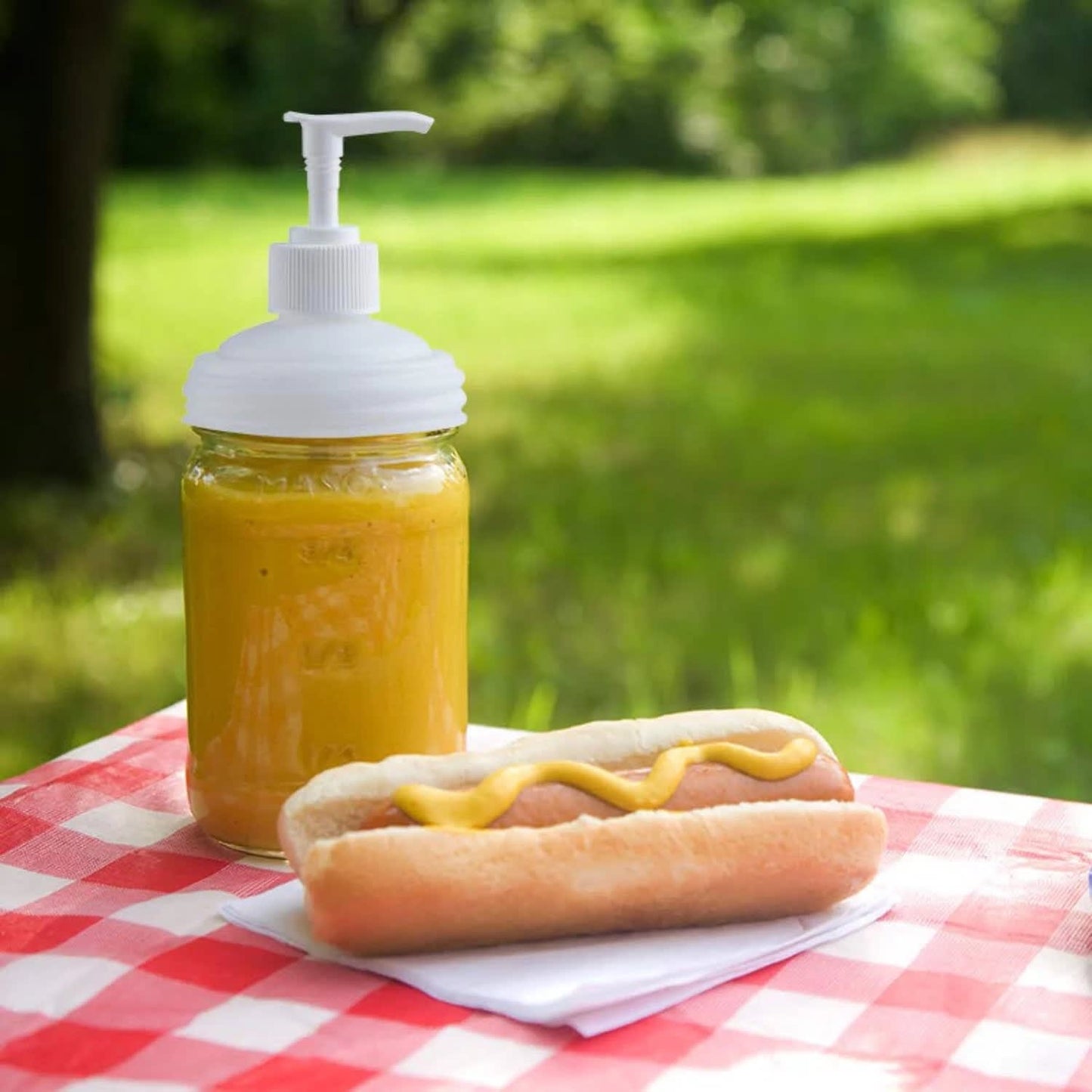 White pump lid fitted on a glass jar filled with mustard next to a hot dog topped with mustard, displayed on a red and white checkered picnic tablecloth, highlighting eco-friendly and USA made Health & Wellness and Home & Decor collections.