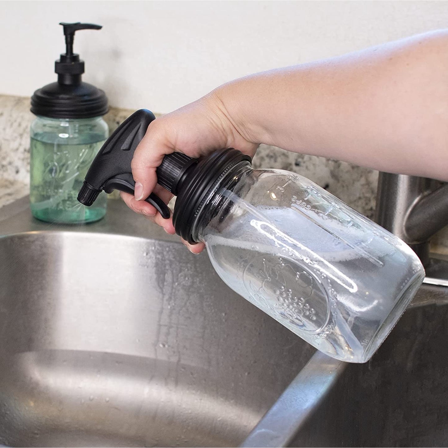 Hand holding a clear regular-mouth glass jar fitted with a black no-break plastic sprayer lid over a kitchen sink, showcasing an eco-friendly home cleaning tool from Household Helpers, USA Made, and Best Sellers collections.