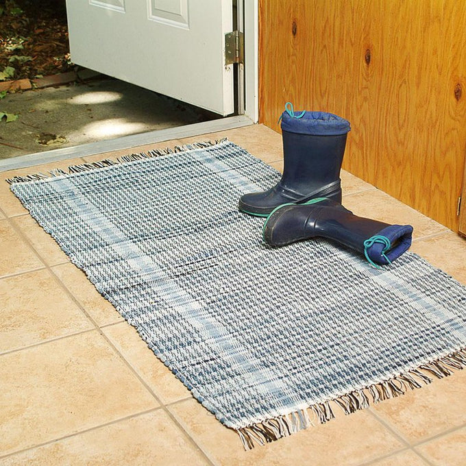 Handwoven denim rug in muted blue shades with fringed edges, placed on a tiled floor near an open door with a pair of navy rain boots, featured in Home & Decor and USA Made collections.