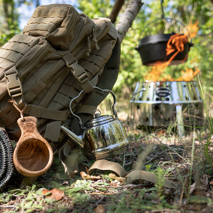 Shiny stainless steel teakettle with a threefold bent handle hanging from a tan backpack on forest floor, beside a wooden cup and camp stove, suited for camping and outdoor collections.