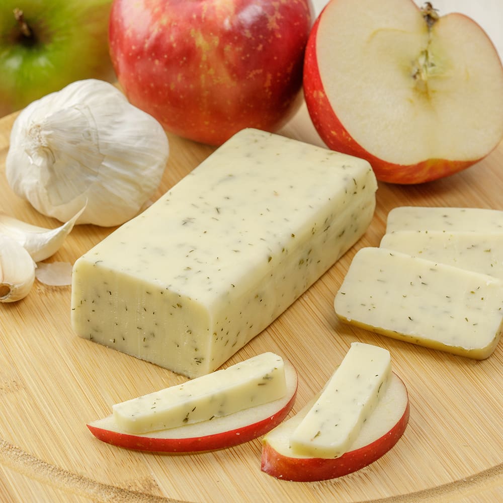Handcrafted block of shelf-stable cheese with herb flecks, sliced and paired with fresh apple wedges and garlic on a wooden cutting board, ideal for Kitchen & Food and Pantry collections.