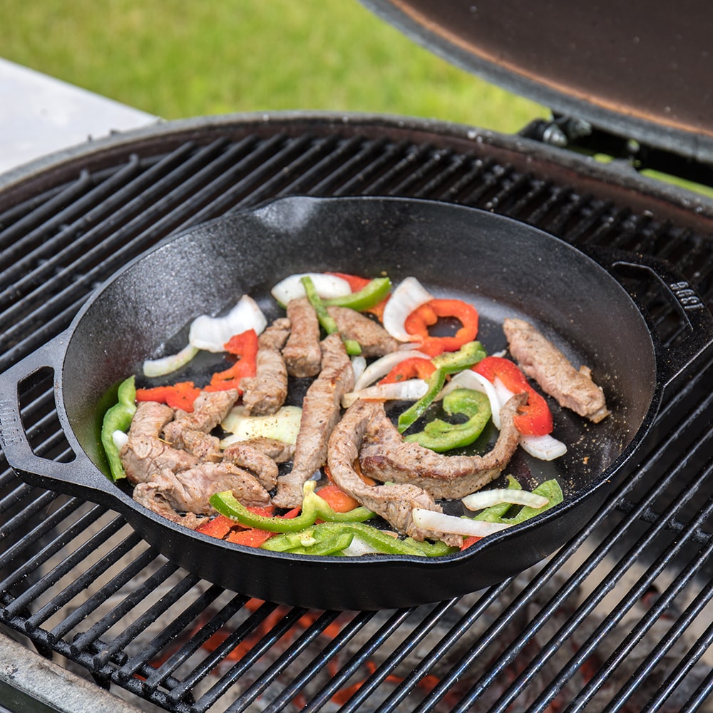 Round cast iron pan with short handles cooking seasoned strips of meat with red and green bell peppers and sliced onions on an outdoor grill, featured in Kitchen & Food, Cookware, Cast Iron, Best Sellers, and Promo Eligible collections.