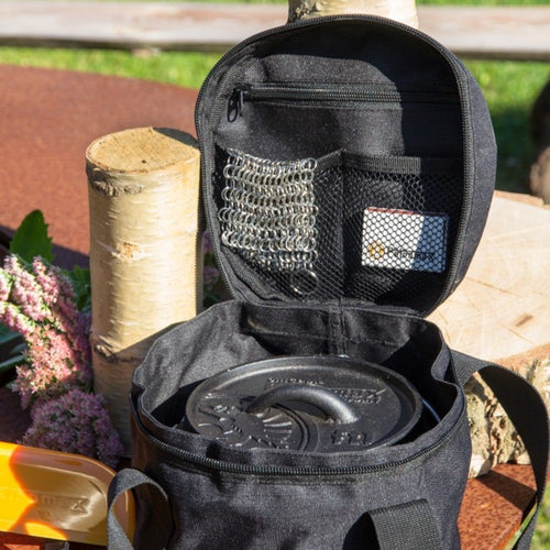 Open black transport bag containing a cast iron Dutch oven with a lid inside, featuring interior mesh pockets holding small accessories, set outdoors with natural wood and flowers, suited for Farm, Garden & Tool, Camping, Best Sellers, and Promo Eligible collections.