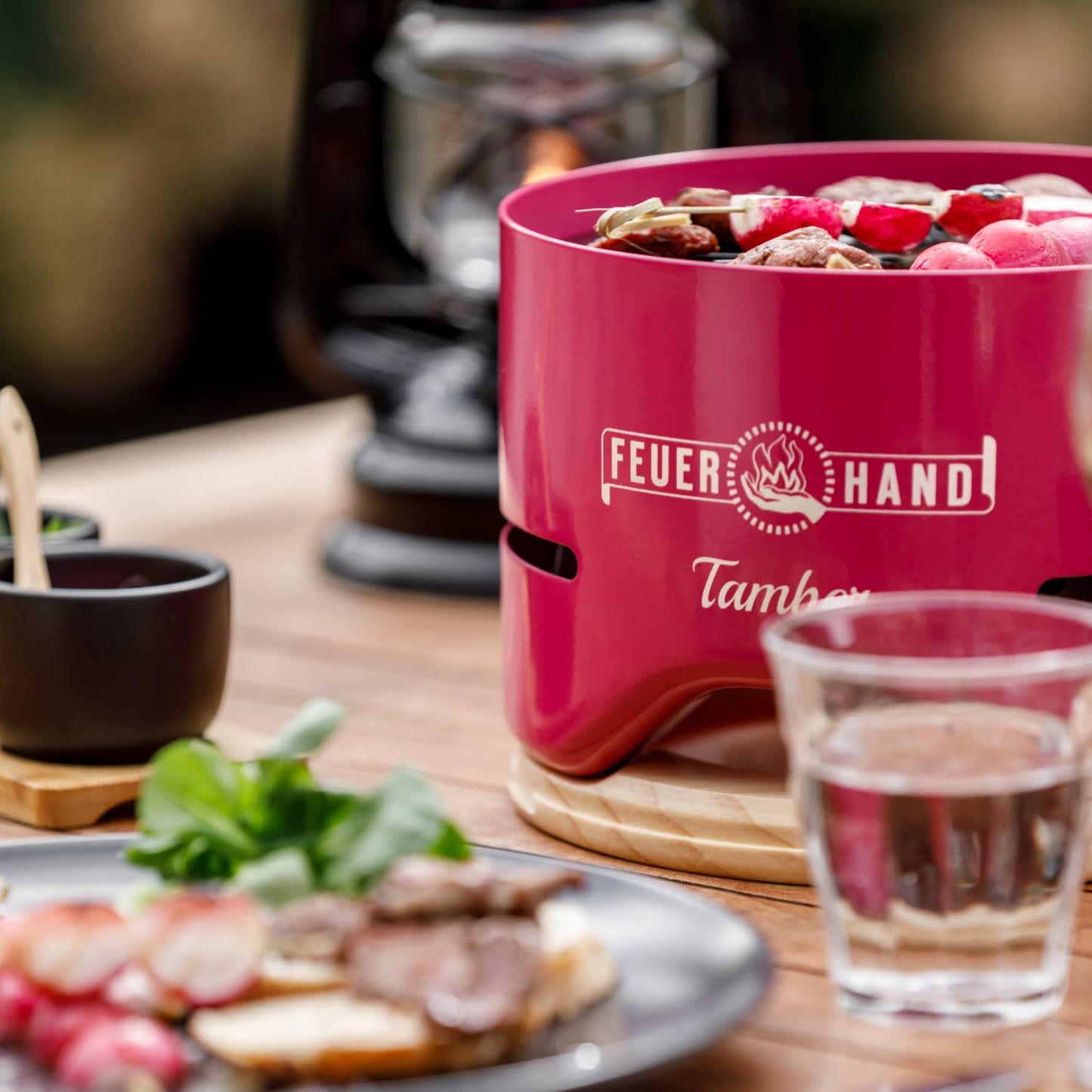 Bright red Feuerhand Tamber tabletop grill with vibrant skewers cooking over low heat, set on a wooden trivet on an outdoor table, featured in Farm, Garden & Tool and Grilling collections.