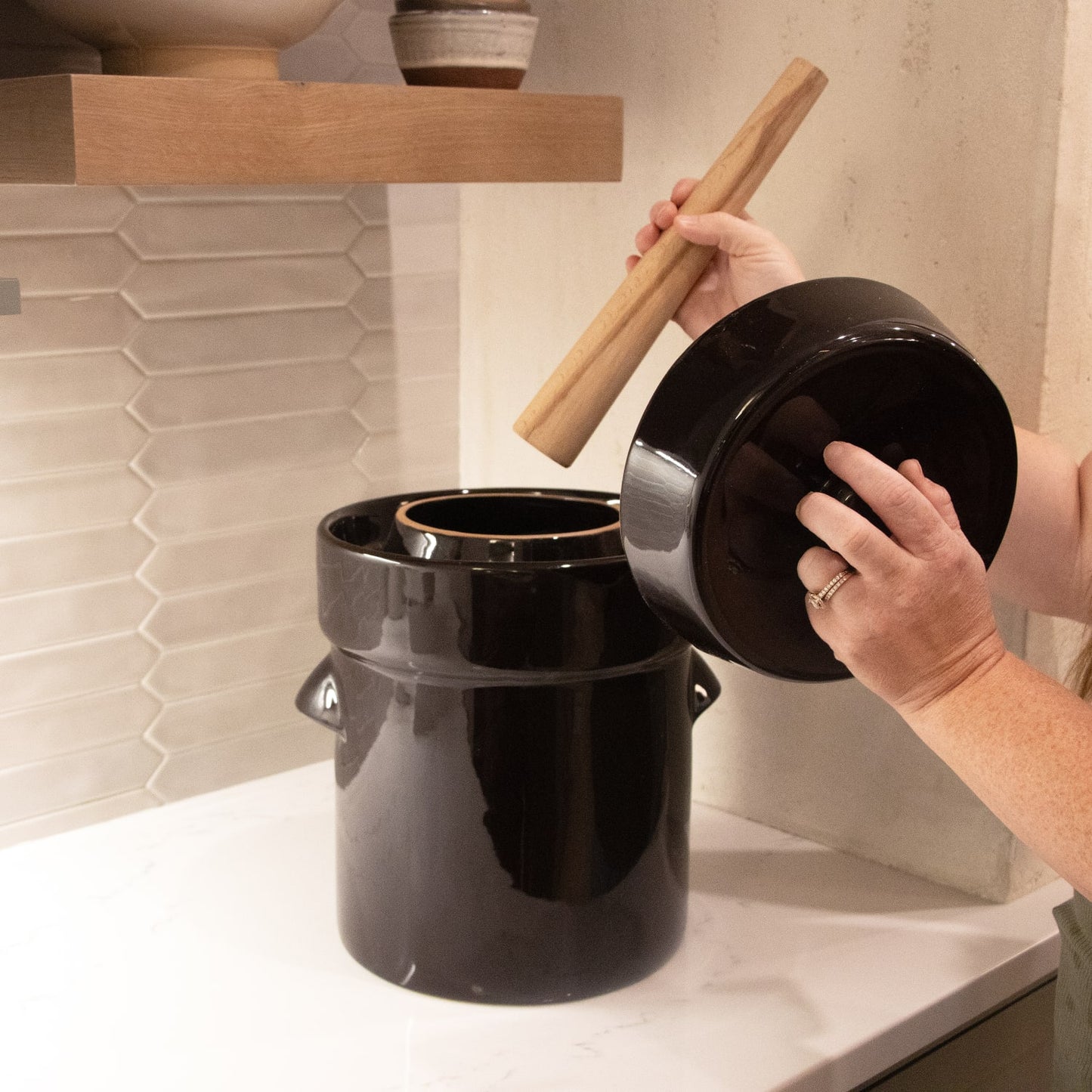 Handcrafted dark stoneware fermenting crock kit with lid and wooden stomper held by hands on a kitchen counter, ideal for Kitchen & Food, Food Preservation, and Fermenting and Pickling collections.