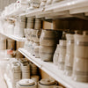 Shelves displaying a collection of handcrafted beige stoneware pitchers, mugs, and covered bowls, each accented with simple stripe patterns, made in USA for Kitchen & Food and Dinnerware collections.