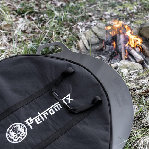 Black durable Petromax carrying bag with sturdy handles protecting a metal griddle near a small outdoor campfire, perfect for camping, farm, garden, and tool use.