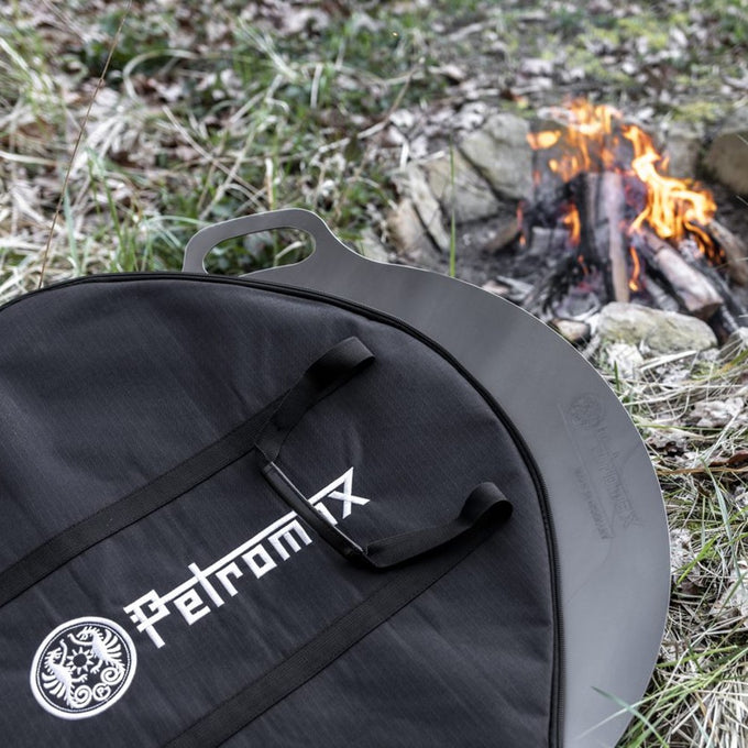 Black durable Petromax carrying bag with sturdy handles protecting a metal griddle near a small outdoor campfire, perfect for camping, farm, garden, and tool use.