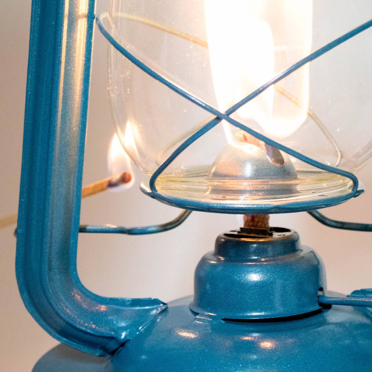 Close-up of a handcrafted blue metal lantern being lit, featuring a clear glass globe with protective wire, ideal for farm, garden, camping, and emergency use.