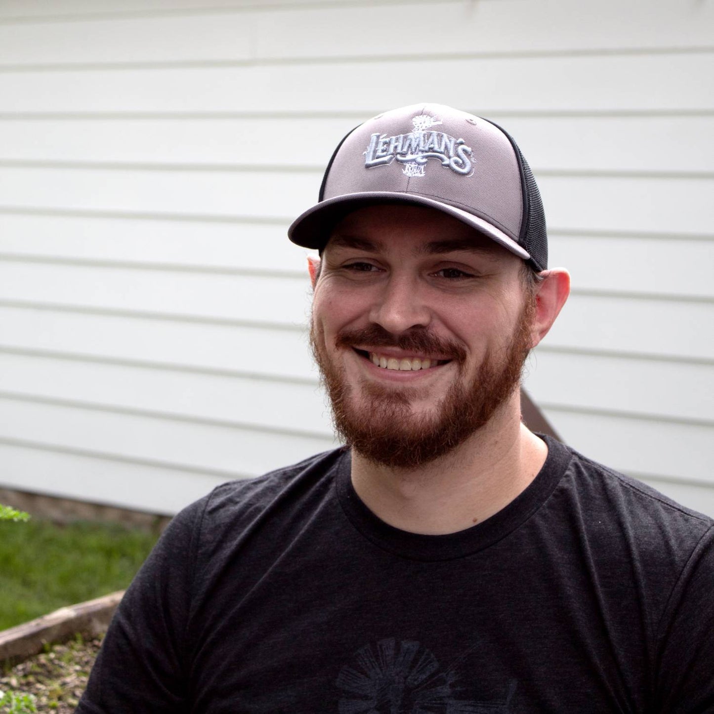 Man smiling and wearing a handcrafted Lehman's embroidered trucker hat with a grey front panel, black stretch mesh back, and slight pre-curved visor, ideal for Health & Wellness and Clothing and Accessories collections.
