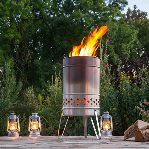 Brushed stainless steel fire barrel with perforated lower venting and sturdy legs, glowing warmly in an outdoor garden setting alongside four lit lanterns, ideal for Farm, Garden & Tool, Camping, and Best Sellers collections.