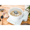 White stand mixer with metal whip driver attachment mixing colorful batter in a bowl on a wooden counter, surrounded by baking ingredients and tools in a kitchen setting