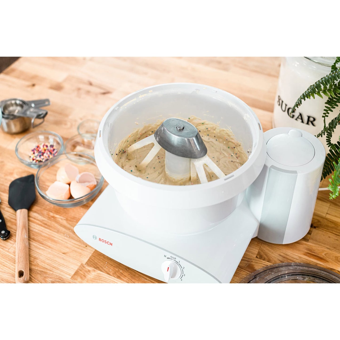 White stand mixer with metal whip driver attachment mixing colorful batter in a bowl on a wooden counter, surrounded by baking ingredients and tools in a kitchen setting