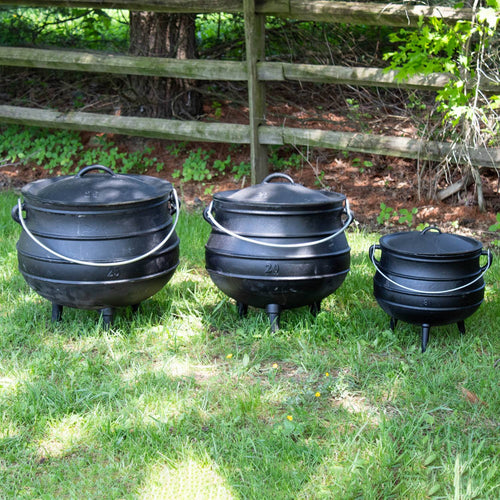 Set of three handcrafted Lehman's cast iron campfire kettles in small, medium, and large sizes with round bodies, fitted lids, wire handles, and sturdy legs, displayed outdoors on grass by a wooden fence in Farm, Garden & Tool, Camping, Best Sellers, and Promo Eligible collections.