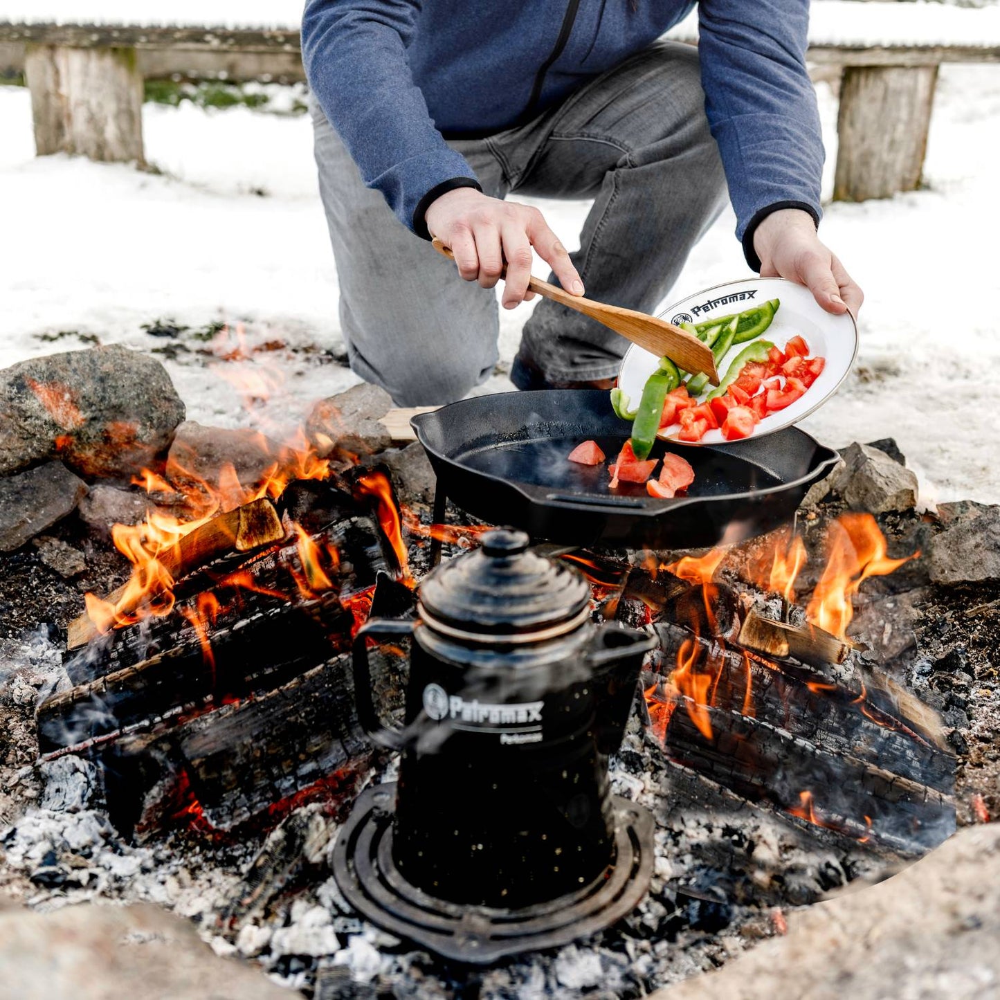 Person using white enamel plate with green and red vegetables to add ingredients into cast iron pan over open campfire, showcasing durable outdoor cookware for camping and kitchen collections.