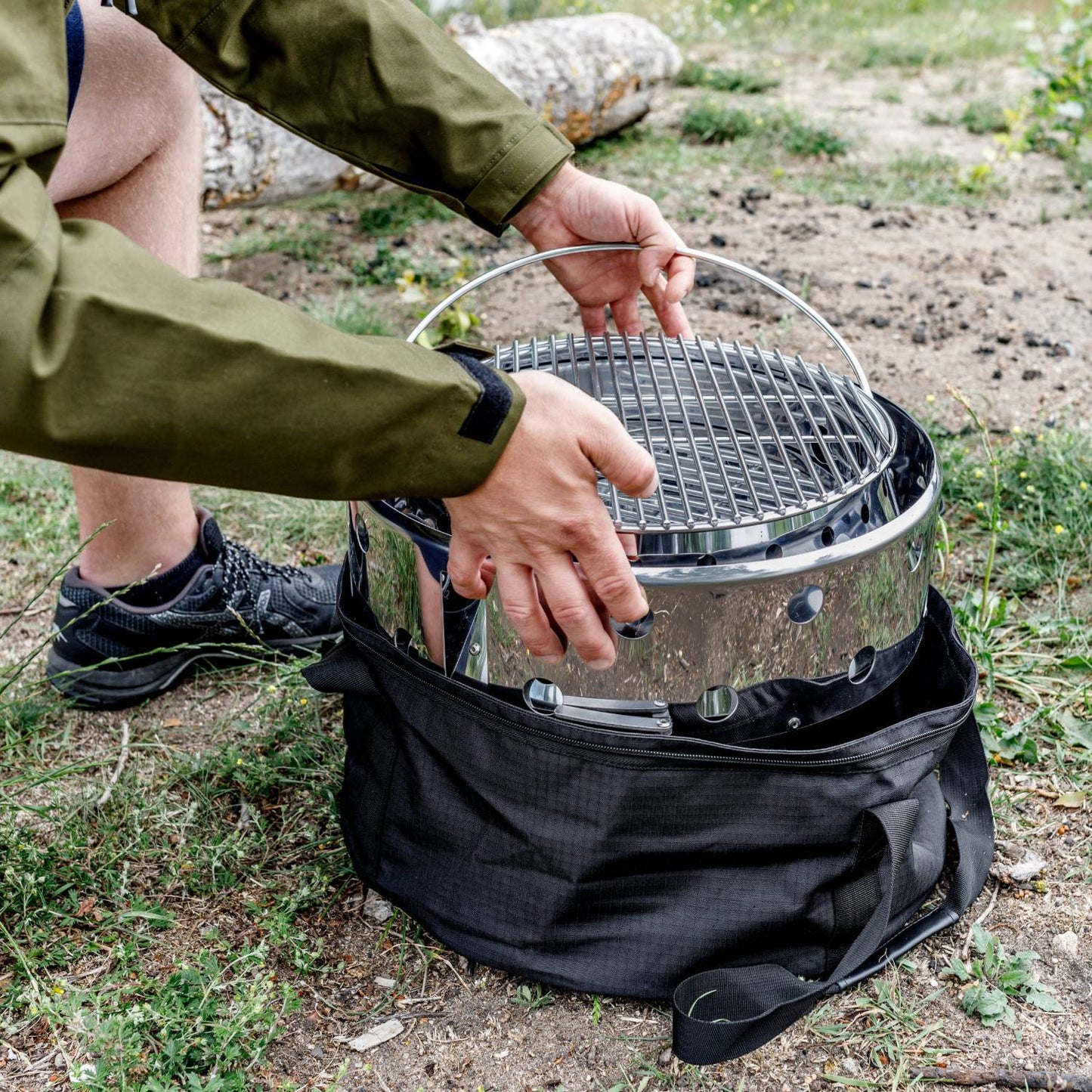 Person placing a portable stainless steel Petromax Atago camp grill and fire bowl into a black carry bag outdoors, designed for versatile use in Farm, Garden & Tool, Camping, and Grilling collections.