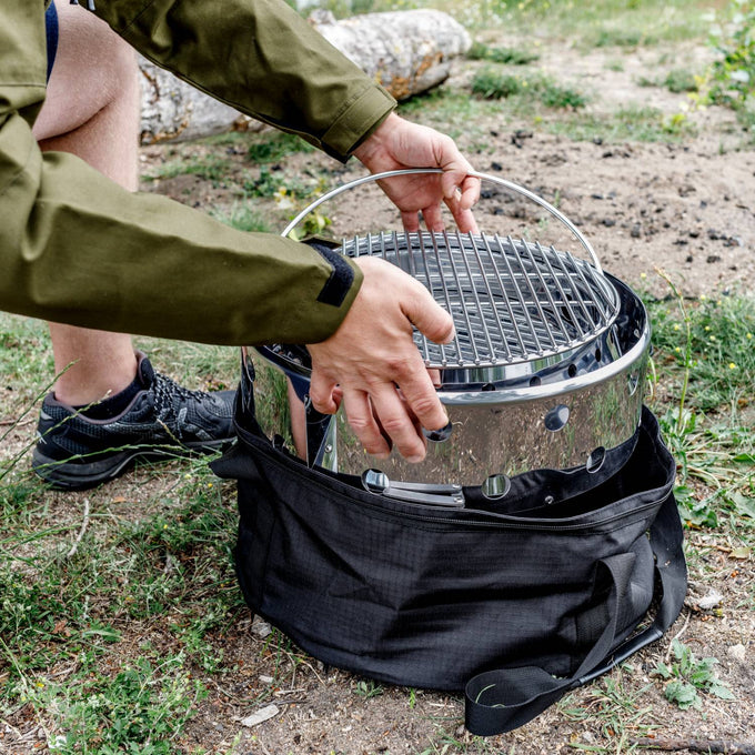 Person placing a portable stainless steel Petromax Atago camp grill and fire bowl into a black carry bag outdoors, designed for versatile use in Farm, Garden & Tool, Camping, and Grilling collections.