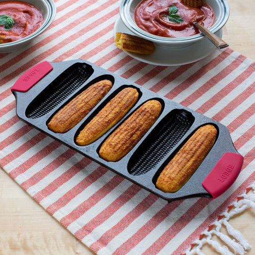 Seasoned Lodge cast iron cornstick pan with six golden cornbread sticks, featuring dual red silicone handles and angled feet, displayed on a striped cloth with bowls of tomato soup, ideal for Kitchen & Food, Cookware, Cast Iron, USA Made, Best Sellers, and Promo Eligible collections.