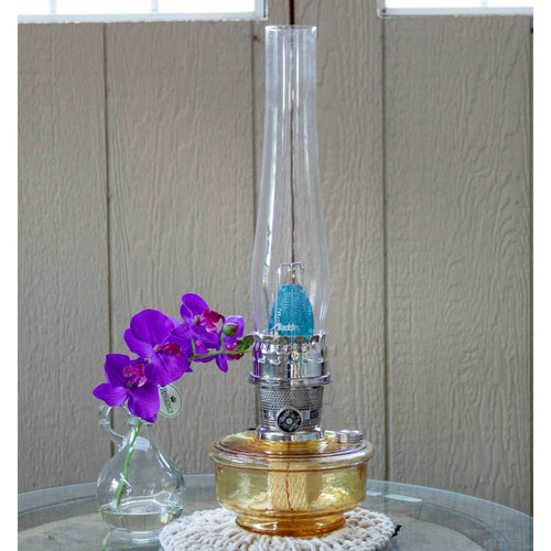 Handcrafted amber glass oil lamp with clear tall chimney and metal burner, displayed on a doily beside a small vase of purple flowers, perfect for Farm, Garden & Tool, Lamps & Lighting, Emergency Supplies, Oil Lamps, Wall Lamps, and Best Sellers collections.