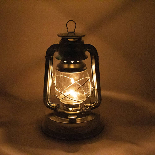 Handcrafted metal oil lantern with glowing flame inside clear glass, casting warm light, ideal for use in camping, farm, garden, pest control, and emergency supplies collections.