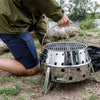 Person adjusting portable stainless steel camp grill with perforated body and foldable legs for outdoor cooking and fires, ideal for camping and grilling in Farm, Garden & Tool collections