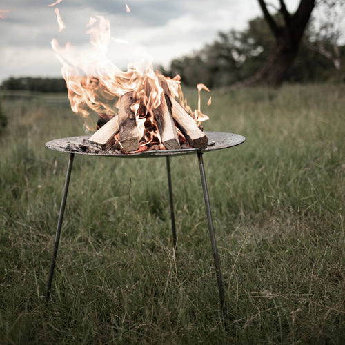 Steel fire bowl with three legs holding a lively wood fire outdoors in a grassy field, perfect for evening campfire gatherings in Farm, Garden & Tool, Camping, Best Sellers, and Promo Eligible collections.