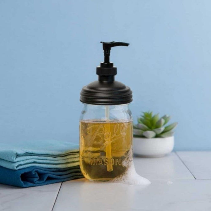 Clear glass mason jar filled with golden soap and topped with a black pump lid, sitting on a white surface with folded blue cloths and a small succulent nearby, highlighting eco-friendly, USA made Household Helpers and Home & Decor collections.